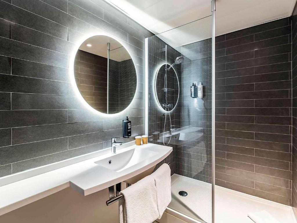 Banheiro do Ibis Lisboa Saldanha com um box de vidro, um espelho redondo iluminado sob uma pia branca com um suporte onde há toalhas brancas penduradas, para representar hotéis baratos em Lisboa