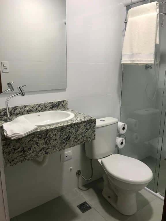 Imagem de um banheiro na Pousada Vila Barboza. Há um espelho, uma pia pequena e um vaso sanitário ao seu lado. Podemos ter uma visão parcial do box.