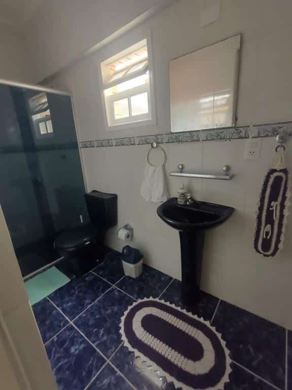 Banheiro com parede de azulejo branco, chão de azulejo preto marmorizado, box preto no lado esquerdo, vaso sanitário preto ao lado do box e pia com pequeno espelho no lado direito do banheiro.