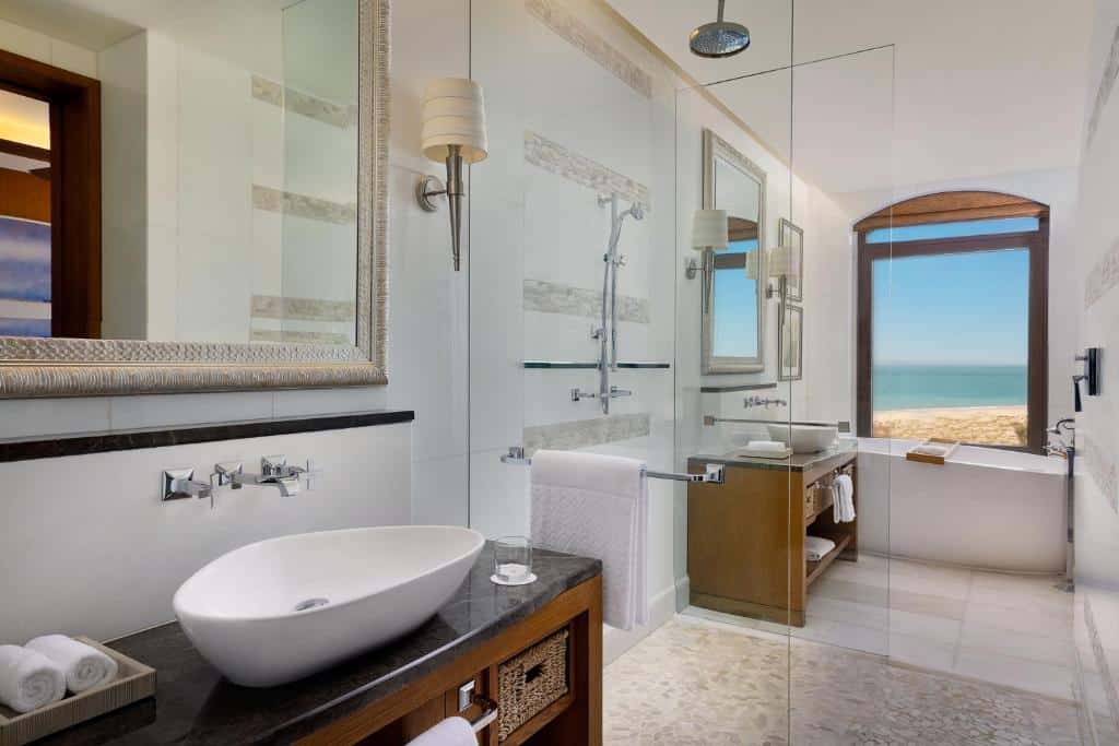 Quarto do The St. Regis Saadiyat Island Resort, Abu Dhabi. A pia no lado esquerdo com um espelho em cima. Uma parede de vidro com uma toalha pendurada, o chuveiro e outra parede de vidro. No fundo, outra pia, uma banheira e uma janela com vista para o mar.
