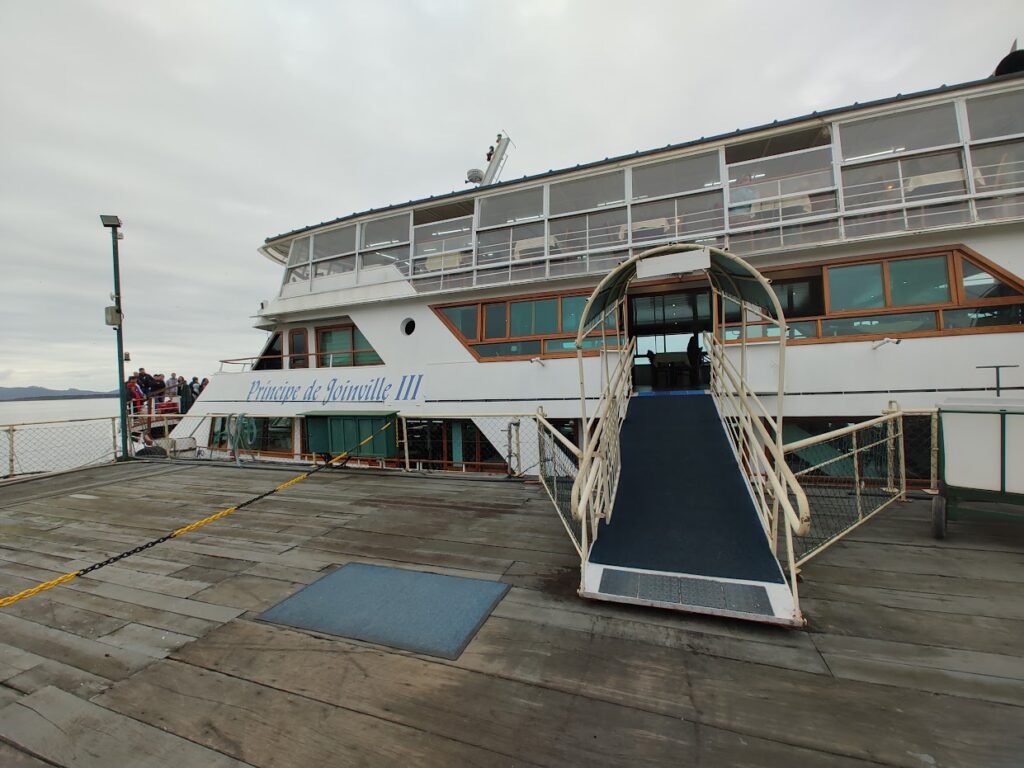 deque de madeira com acesso coberto com rampa para o Barco Príncipe. O barco é branco e possui três andares com muitas janelas.