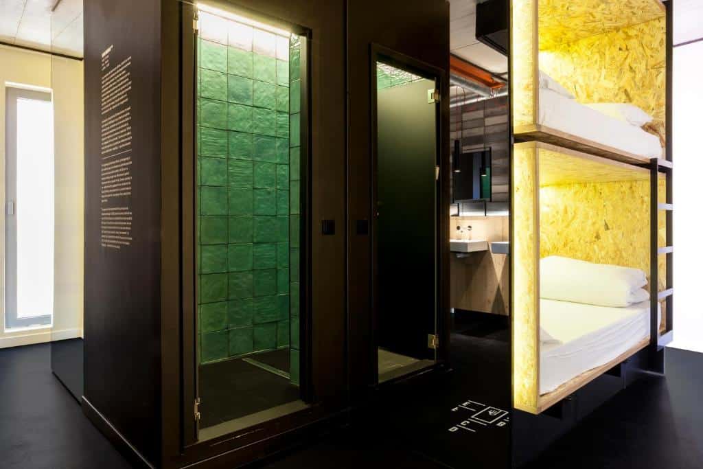 Quarto do Bastardo Hostel, uma das recomendações de hostels em Madri. Duas camas em beliche estão no lado direito da imagem, e um espelho reflete a imagem da pia no banheiro. Uma porta leva a um lugar com azulejos verdes.
