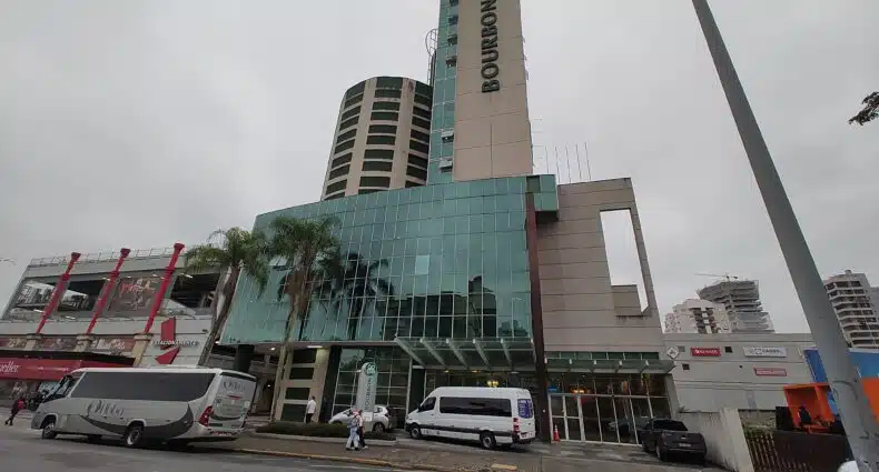 fachada do hotel bourbon joinville convention onde é possível enxergar a rua com alguns carros passando em frente, e duas torres no prédio, uma menor do que a outra, sendo a menor arredondada.