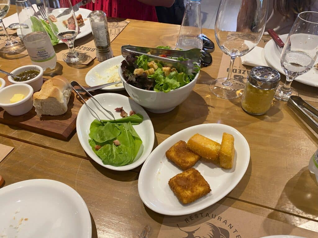 Pratos de acompanhamentos do restaurante BraZedo, com tábua de pão e manteiga e geleia, saladas, ou polenta e queijo fritos