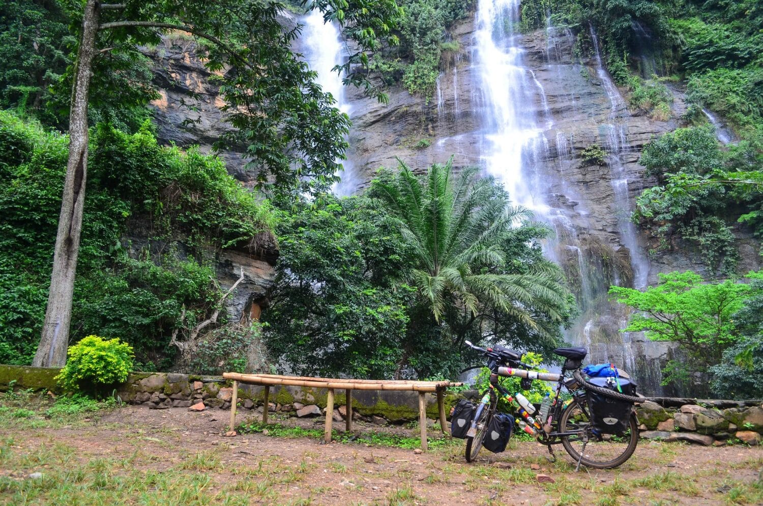 Cachoeira Kpime durante o dia com queda d'água ao fundo, no meio das rochas, em volta vegetação e na frente da cachoeira uma bicicleta parada no lado direito da imagem. Representa seguro viagem Togo.