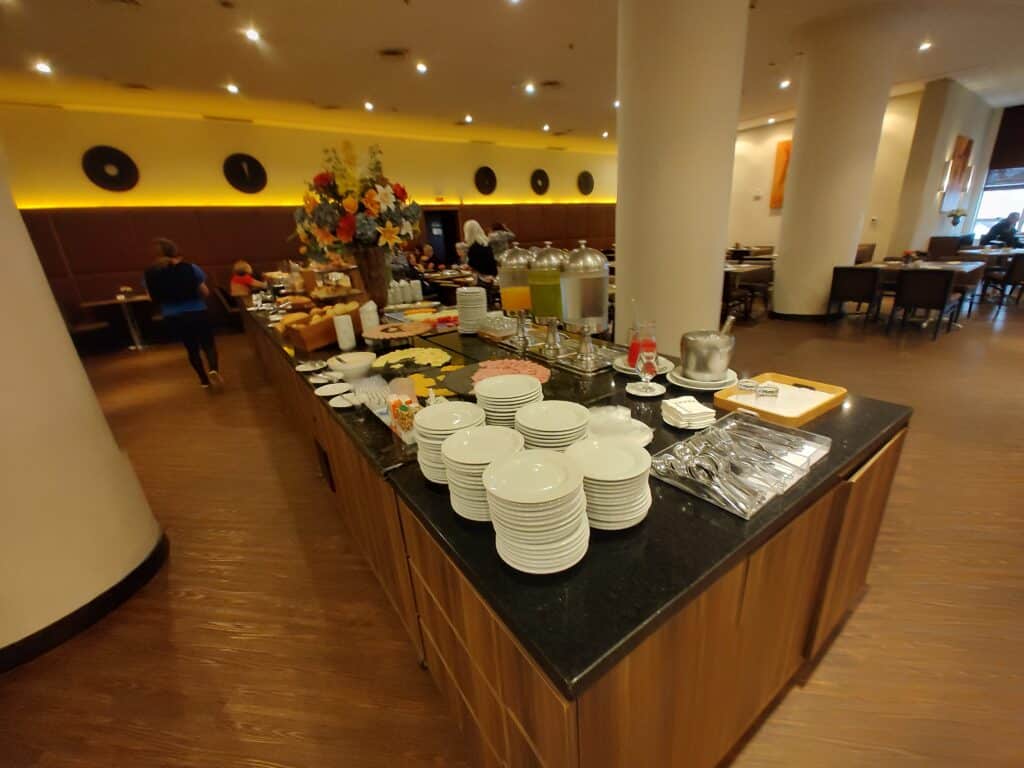 Refeitório do Bourbon Joinville Convention Hotel mostrando a mesa de buffet com vários pratos e louças, sucos, frios, pães e frutas.