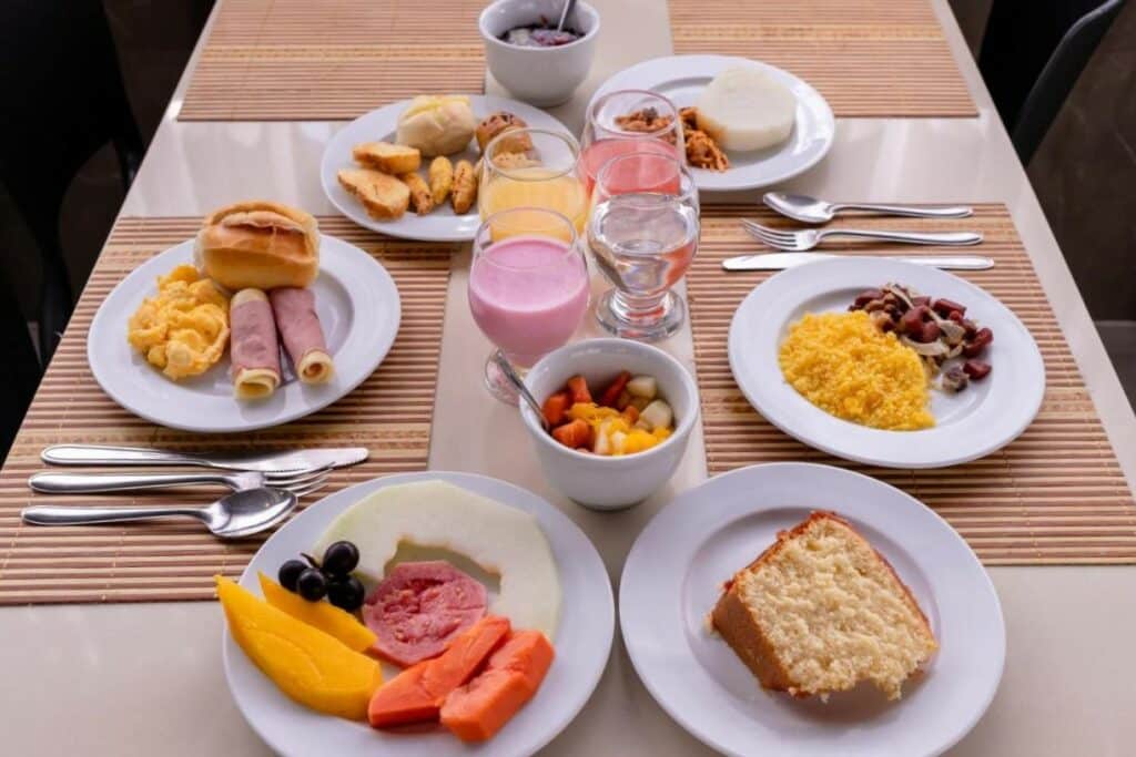 Mesa com café da manhã com pratos de frutas, bolo, pães, ovos, cuscuz, salada de frutas, copos com suco, iogurte e água. Foto para ilustrar post sobre pousadas em Campina Grande.