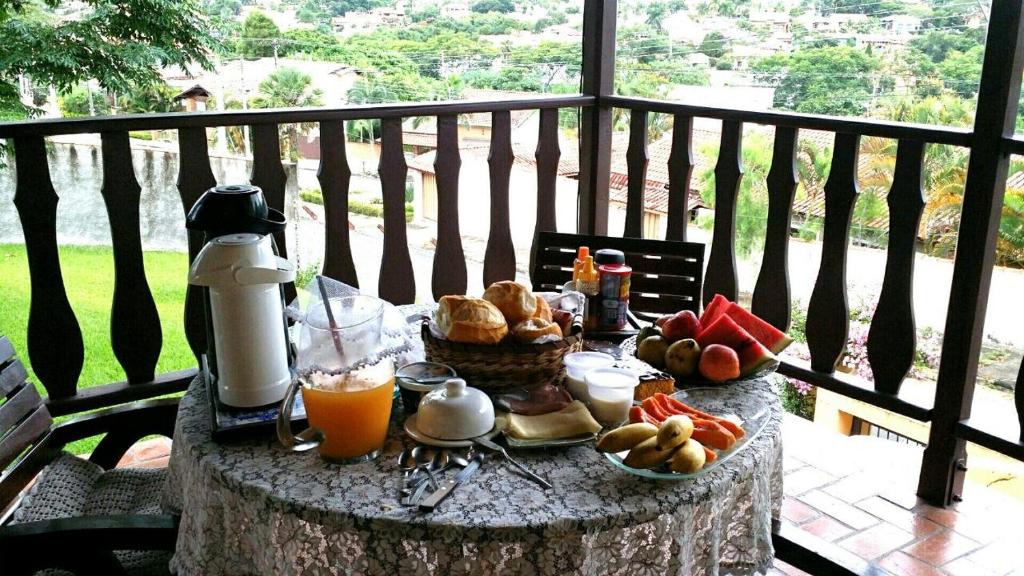 Uma mesa redonda com vários alimentos de café da manhã na Pousada Suítes Águas de São Pedro. A mesa está situada em uma varanda, e possui suco, café, pães, frios e frutas.