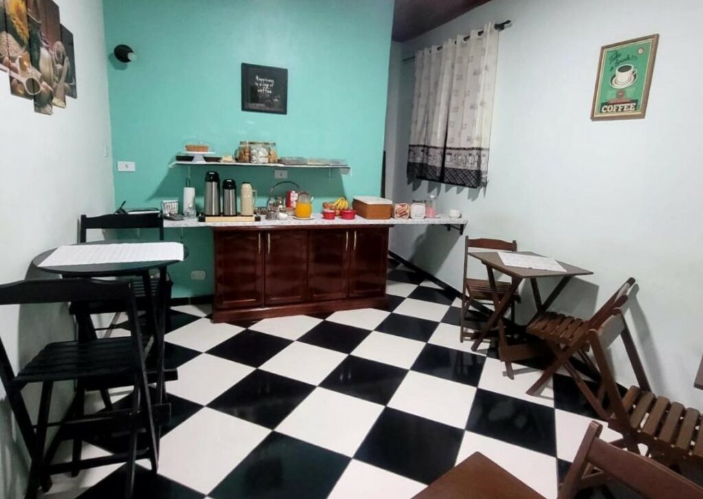 Espaço do Hotel Serra do Mar para tomar café. O chão com piso quadriculado, mesas de madeira com duas cadeiras. Atrás uma mesa com frutas, café e bolachas.