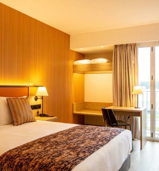 Quarto do Hotel 3K Porto Aeroporto, com cama de casal do lado esquerdo, com uma luminária do lado esquerdo da cama com uma mesa com cadeira próximo. Representa hotéis perto do aeroporto no Porto.