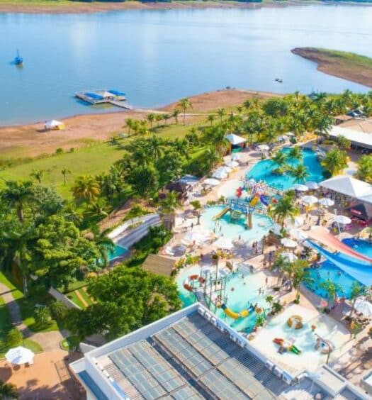 Foto aérea de um pedaço amplo com várias piscinas com água azul, algumas construções em volta, várias árvores e um longo gramado verde perto do rio, ilustrand post Hotéis em Caldas Novas.