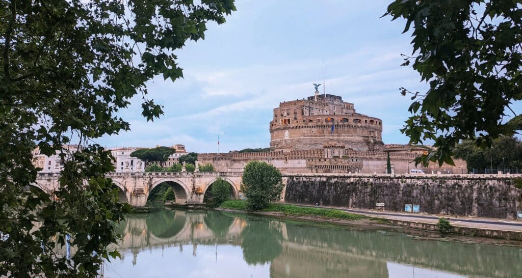 vista da ponte e do Castelo de Santo Ângelo, em Roma, uma fortaleza de pedra acima de um rio, com esculturas de anjos por todo o caminho