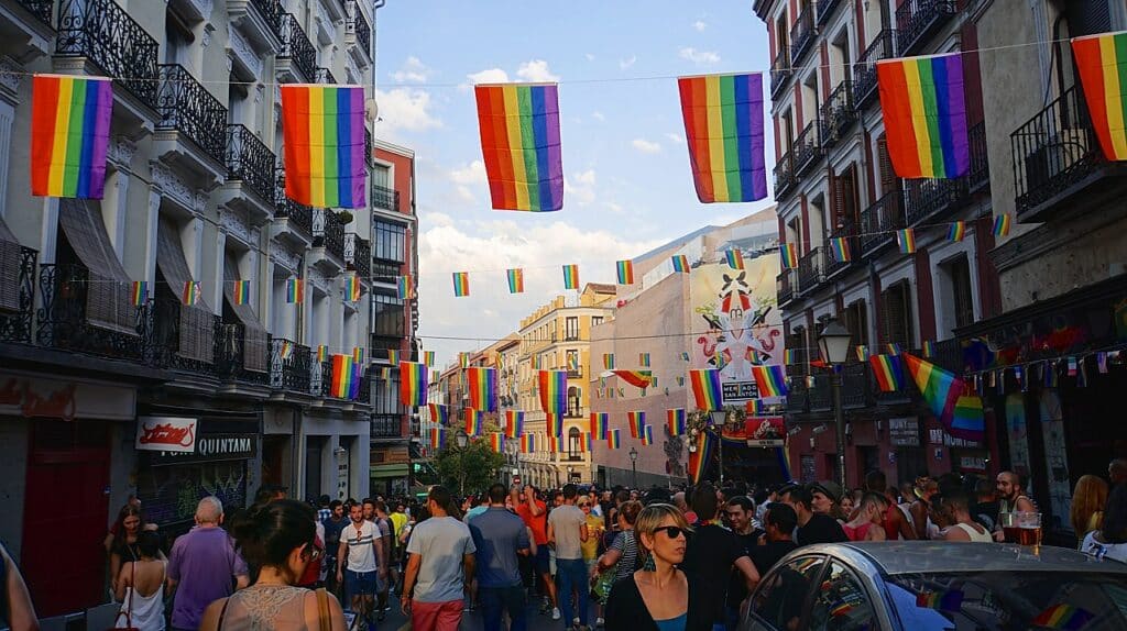 Bandeiras do arco-íris penduradas acima de uma rua no bairro de Chueca. Várias pessoas caminham por ali. - Foto: Ted Eytan via Wikimedia Commons