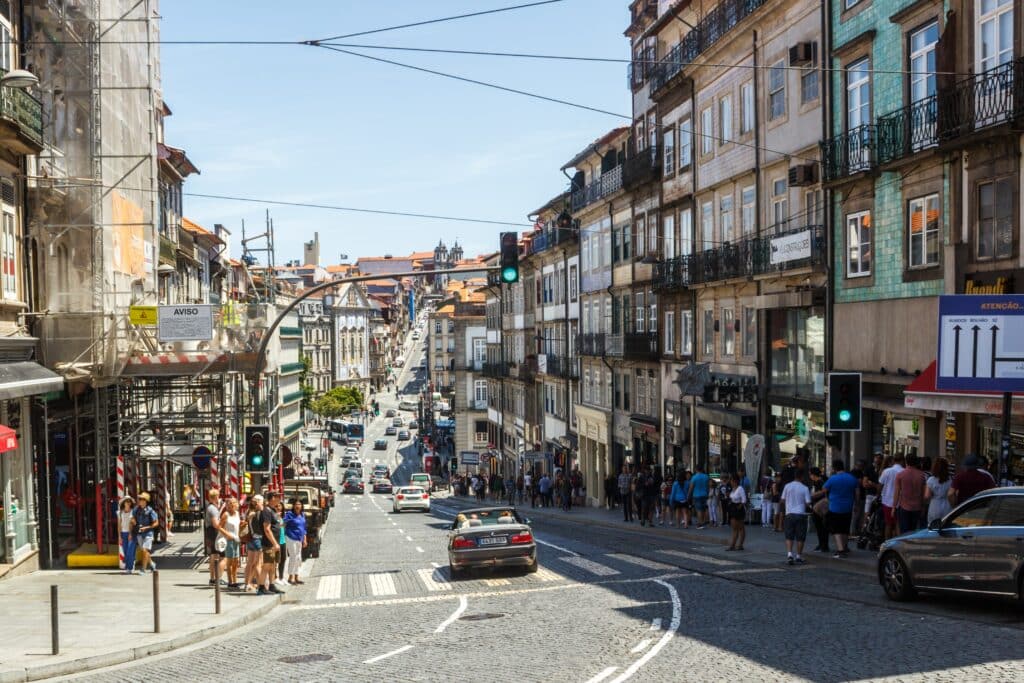 Rua no Porto, Portugal durante o dia cm carros circulando na rua e em cada lado da rua calçadas cheias de pessoas.