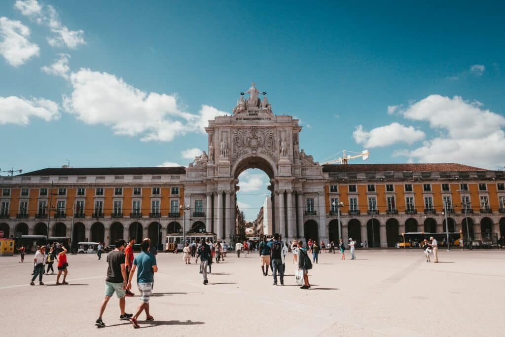 Praça do Comércio em Lisboa com um grande arco com esculturas, o prédio é de época em tons de cinza e laranja, há muitas pessoas caminhando pelo local