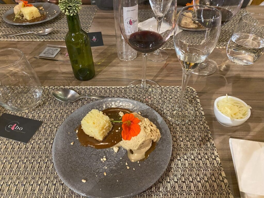 Prato de sobremesa no Cobo Wine bar, onde se vê um pedaço de bolo simples sobre uma calda de caramelo, ao lado de uma bola de sorvete de tom amadeirado, com uma flor por cima. Nos arredores do prato há taças com vinho branco e tinto, além de um copo de água