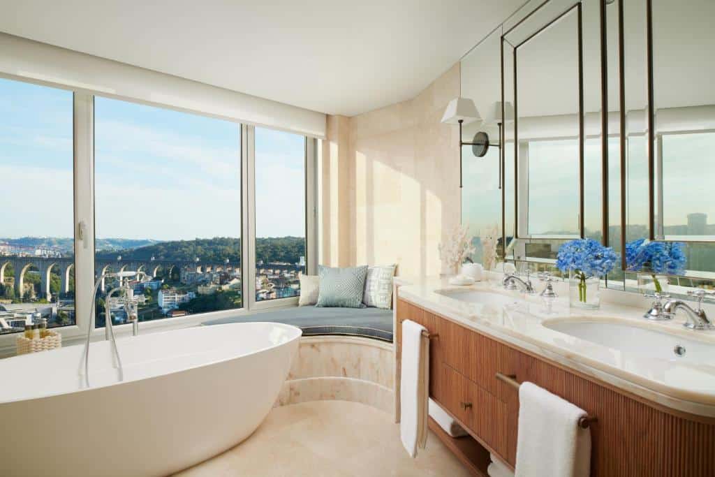Banheiro do Corinthia Lisbon com uma janela enorme com vista para a cidade, há inclusive um pequeno sofá no canto perto da janela, além de uma banheira oval e uma pia com duas cubas e um amplo espelho, para representar hotéis para brasileiros em Lisboa