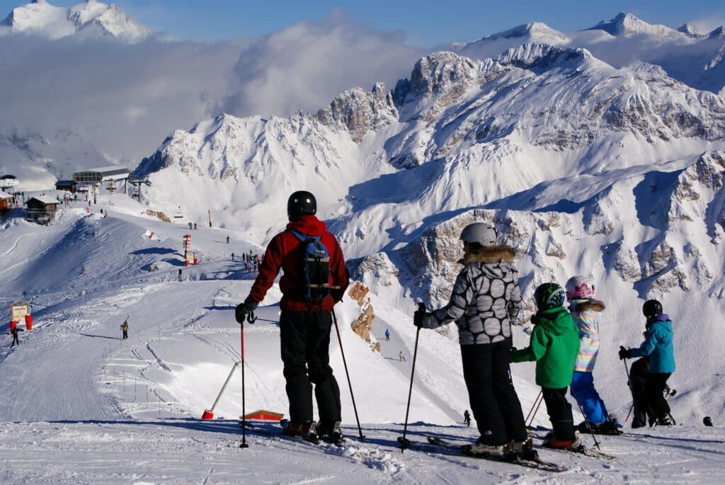 Grupo de pessoas em roupas de frio e com equipamentos de esqui na neve olhando para frente. Todos estão com capacetes. A paisagem na frente deles consiste em montanhas cobertas de neve, algumas propriedades ao fundo e pessoas andando.