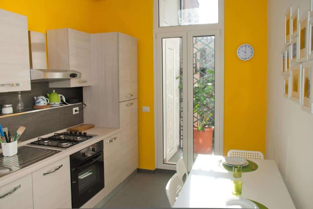 cozinha do Welcome Friends completa e equipada, com armários, fogão e geladeira, mesa com cadeiras brancas com paredes amarela e uma janela grande que tem vista