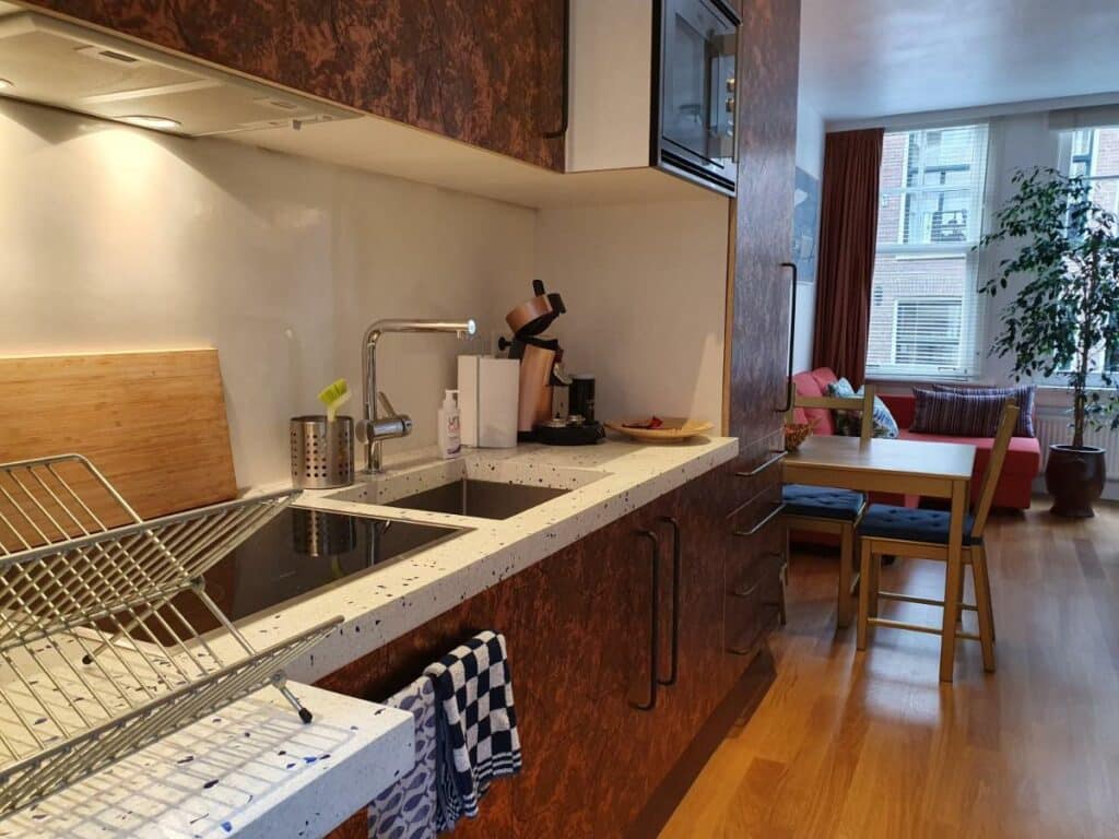 Cozinha do Huis Roomolen, com uma pia no lado esquedo, com escorredor de louças de metal, tábua de madeira e máquina de café em cima. Há, também, acima da pia uma ármário suspenso com forno e, ao fundo do ambiente, uma mesa de madeira, sofá vermelho, um vaso de flor e uma janela iluminando o local com luz natural