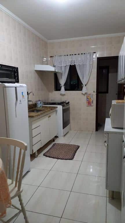 Cozinha com pequena janela com cortina e porta na parede aos fundos, fogão, pia e geladeira no lado esquerdo e armários com microondas no lado direito, no Apartamento Pé na areia Mongaguá.