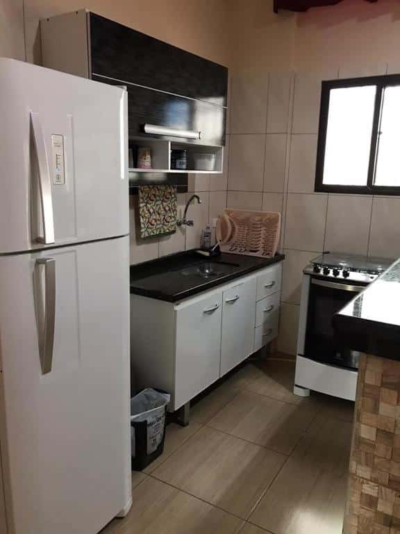 Pequena cozinha com geladeira no lado esquerdo, pia com armário em cima ao lado da geladeira, fogão e janela ao lado direito.