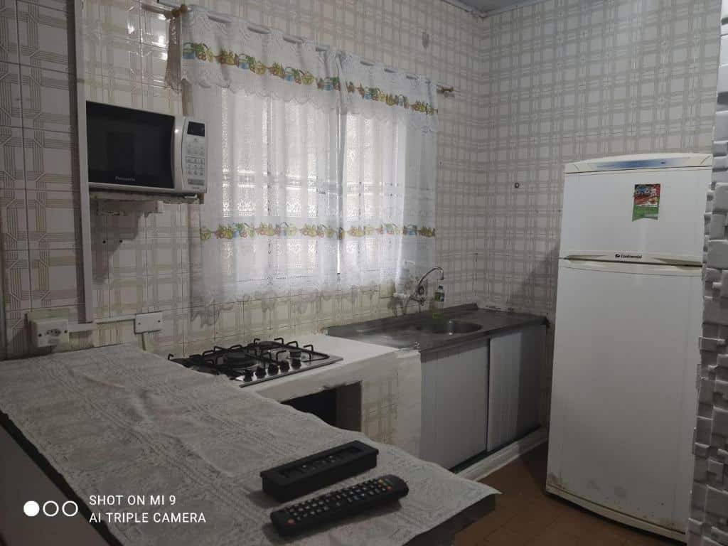 Pequena cozinha com bancada, fogão e microondas ao lado esquerdo, pia, janela com cortina e geladeira no lado direito.