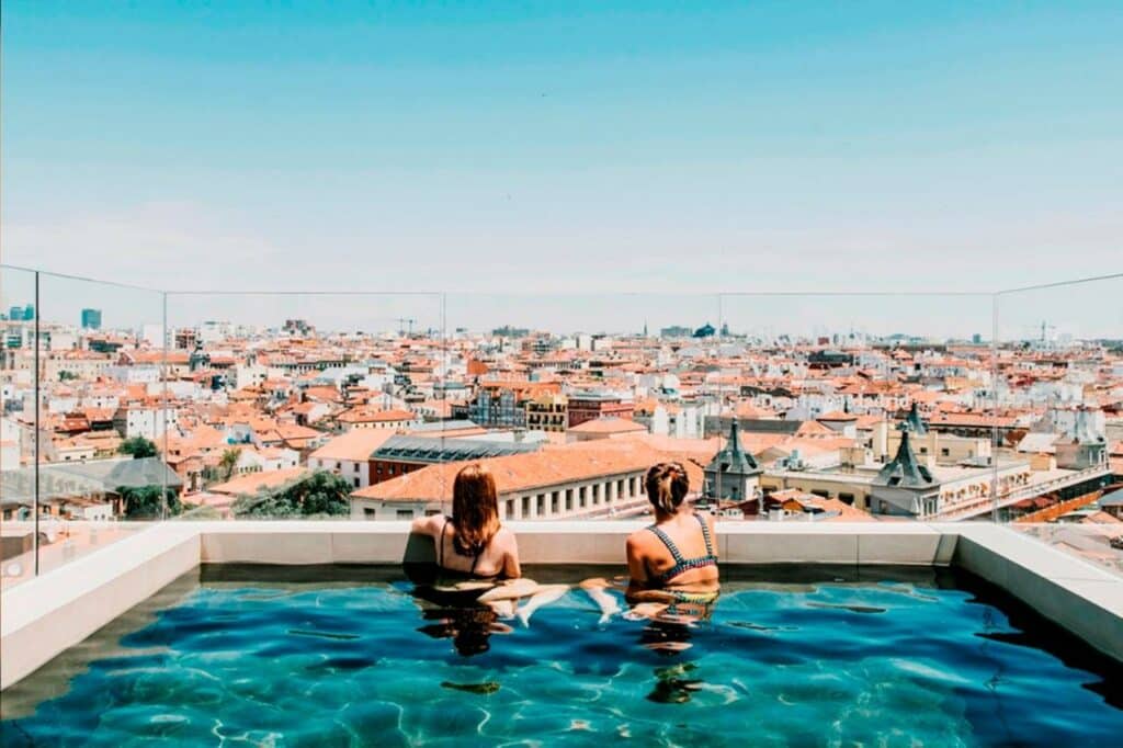 Duas mulheres sentadas dentro da piscina do Dear Hotel Madrid observando a vista da cidade. As duas usam bíquini e estão de costas para a foto, apenas com a parte de cima do corpo para fora d'água. A foto foi tirada de dia e há proteção de vidro ao redor da piscina.