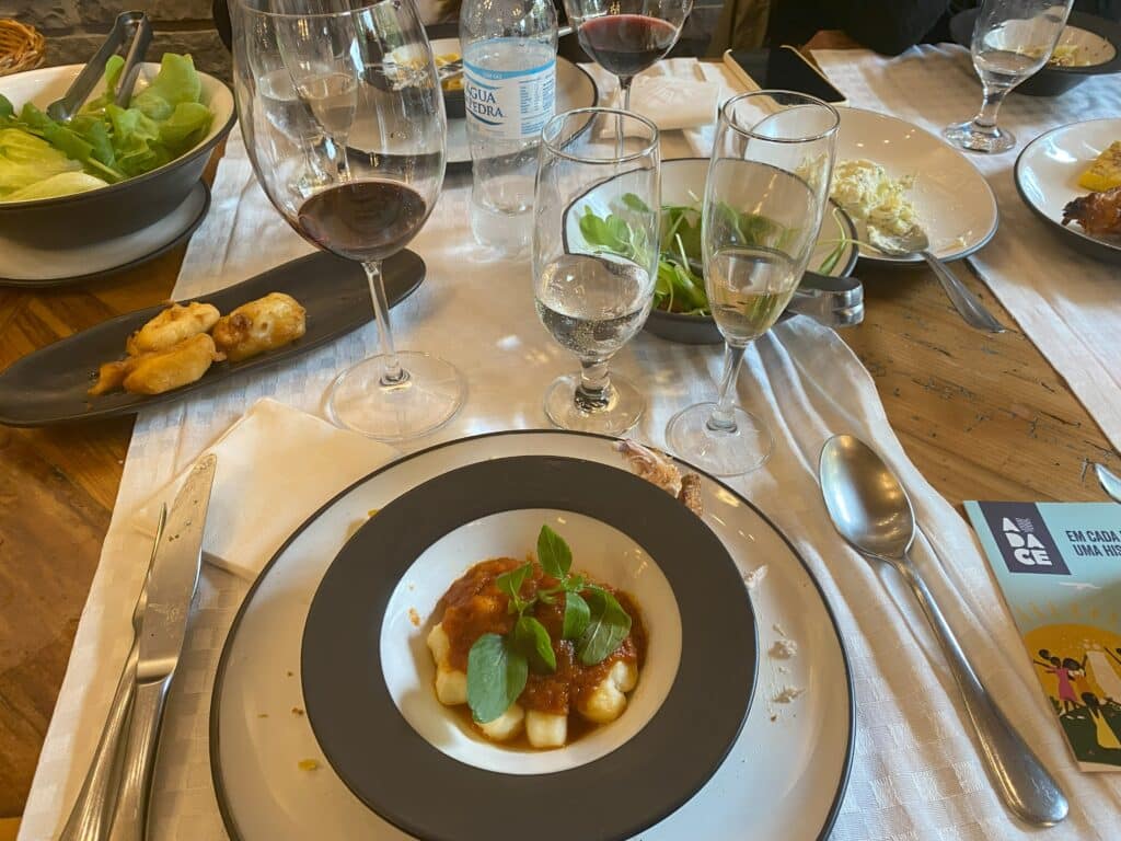 Foto de pratinho fundo, com nhoque ao molho sugo, servido no restaurante Di Paolo. Atrás do prato há três taças, uma com vinho tinto, uma com água e uma com vinho branco