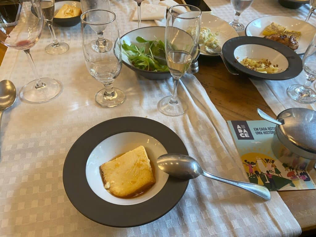 Mesa do restaurante Di Paolo, com salada e demais acompanhamentos (maionese, macarrão, galeto e polenta) ao fundo, e em primeiro plano um prato com pudim