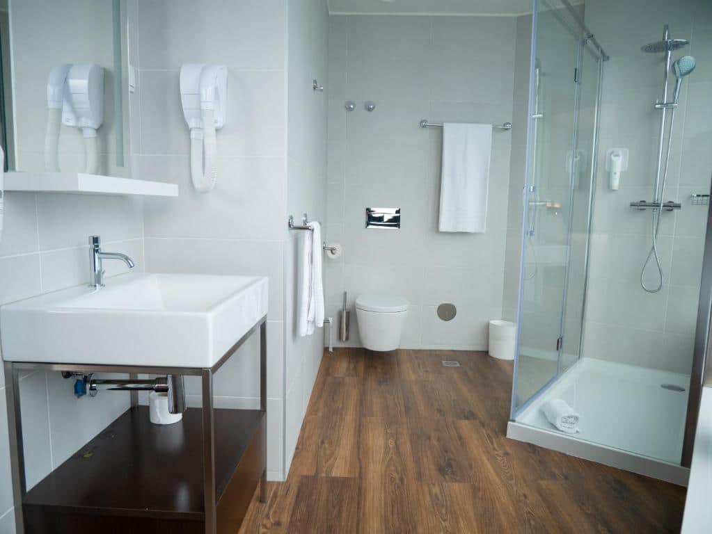 Banheiro do easyHotel Lisbon, do lado direito há um box, do lado esquerdo uma pia com um móvel embaixo, há também um espelho e pelo banheiro há toalhas brancas penduradas