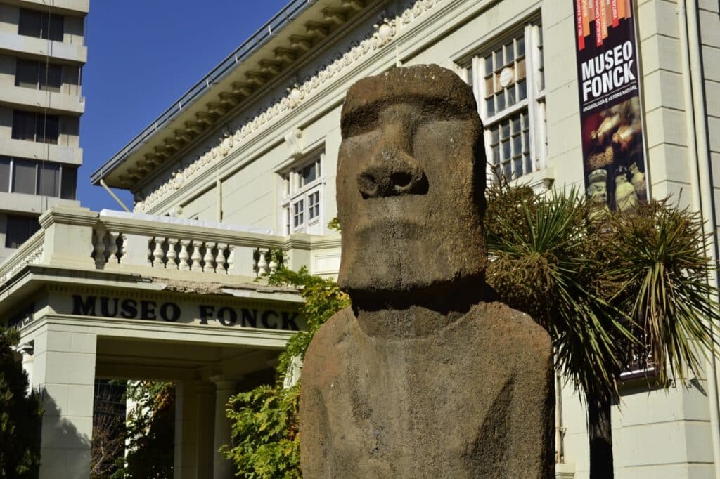 Foto da estátua Moai no Museu Fonck. Ela é bem grande e está ao ar livre. No fundo está o próprio museu e sua fachada.