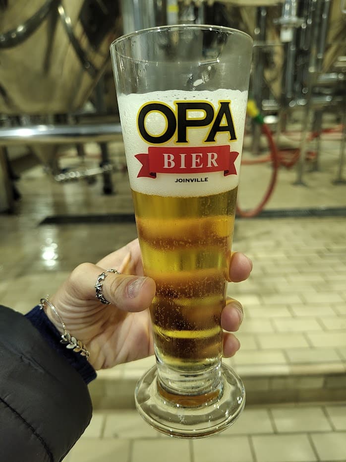 copo de vidro da Opa Bier Joinville com cerveja dentro. Uma mão feminina segura o copo.