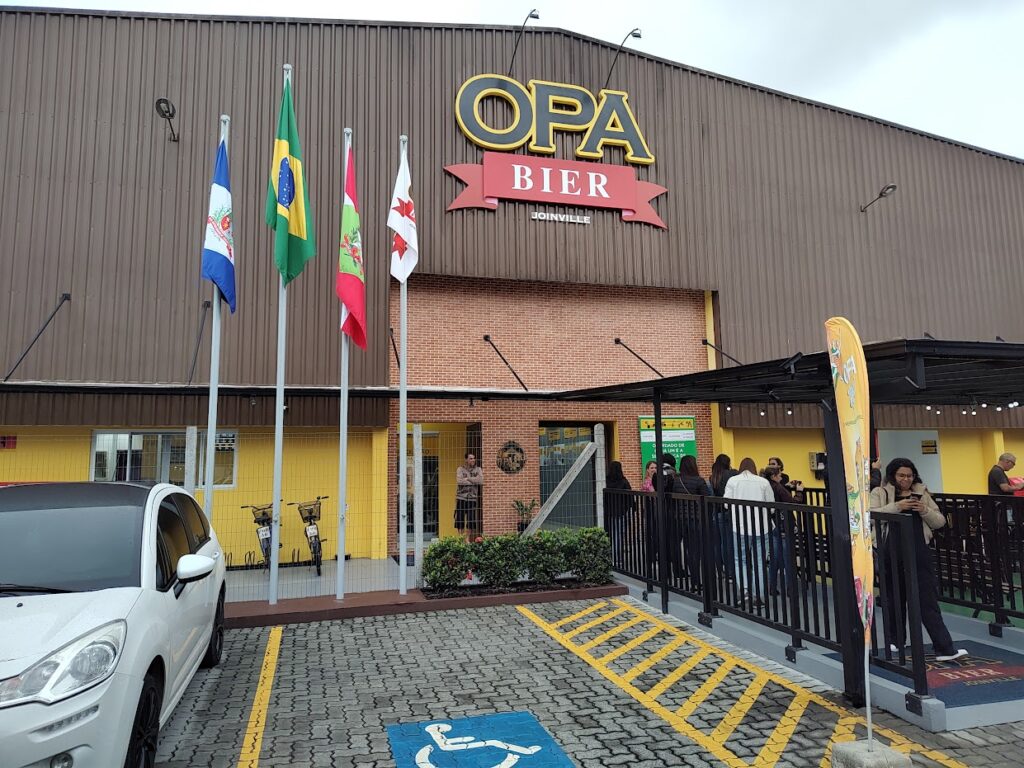 fachada da fábrica de cerveja Opa Bier em Joinville com quatro bandeiras penduradas na entrada, além de um estacionamento e uma fachada de tijolos.