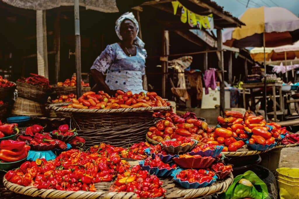 Feira em Abeokuta, Nigéria durante o dia com pimentas e iguarias a frente e ao fundo uma mulher negra.