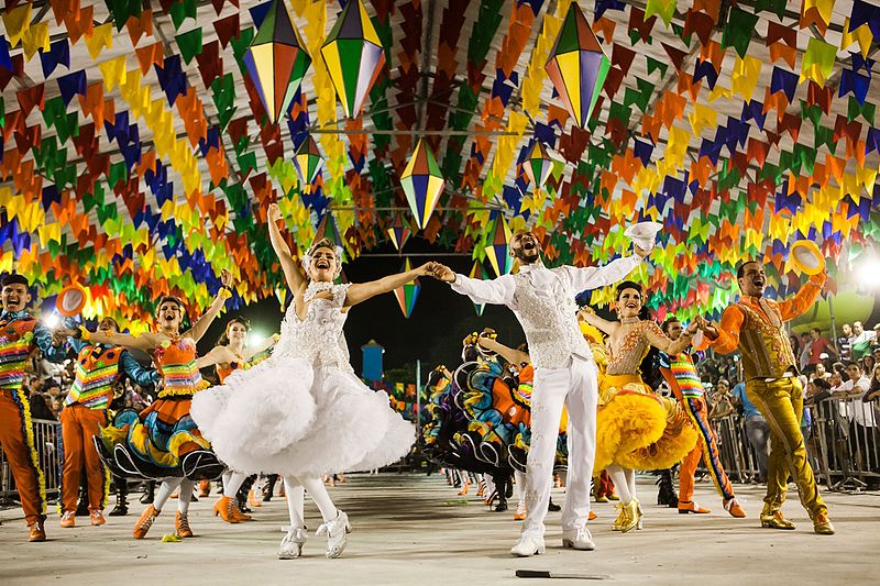 São João de Caruaru com casais de dançarinos enfileirados com roupas coloridas atrás, um casal de dançarinos com roupas brancas em destaque na frente e bandeirinhas e balões de decoração de festa junina no teto.