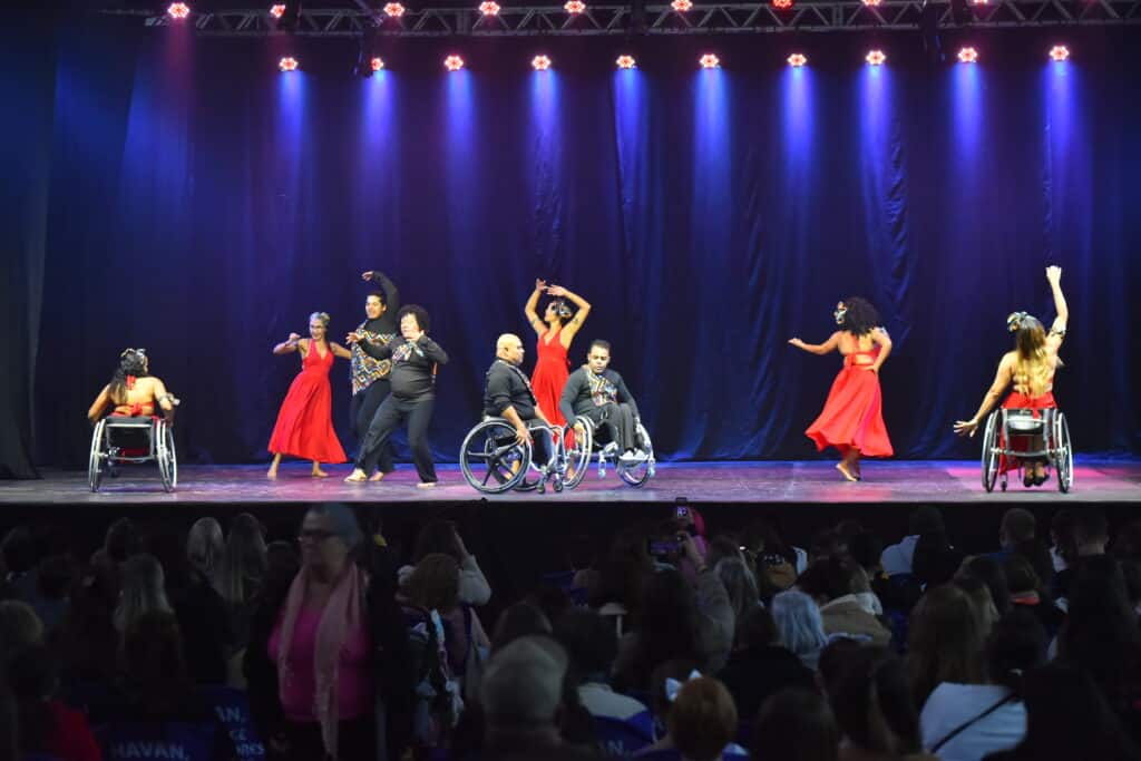 Nove dançarinos, entre homens e mulheres, dançando no palco fechado. Alguns dançam usando cadeiras de roda, outros estão em pé. As mulheres usam um vestido vermelho esvoaçante e os demais usam calça e camiseta de mangra comprida preta.