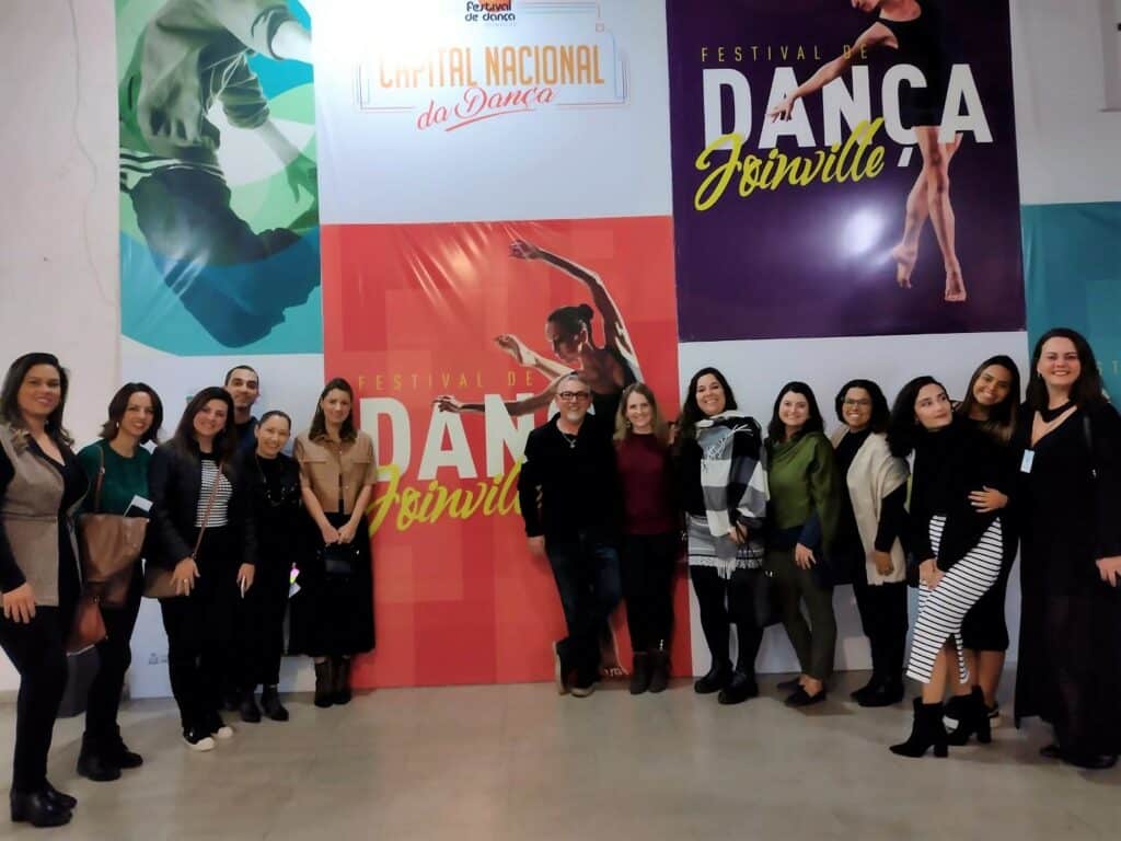painel gigante e colorido com várias imagens retangulares e verticais de mulheres dançando, representando diferentes modalidades de dança. Em frente há um grupo de mulheres posando para a foto.