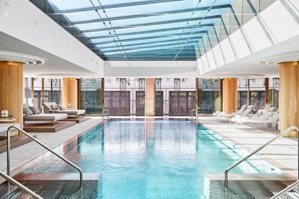 Área da piscina do Four Seasons Hotel Madrid. O local é amplo e com teto de vidro. Há espreguiçadeiras e almofadas dos dois lados da piscina, que está no centro do lugar e tem escadas nas quatro pontas com corrimão.