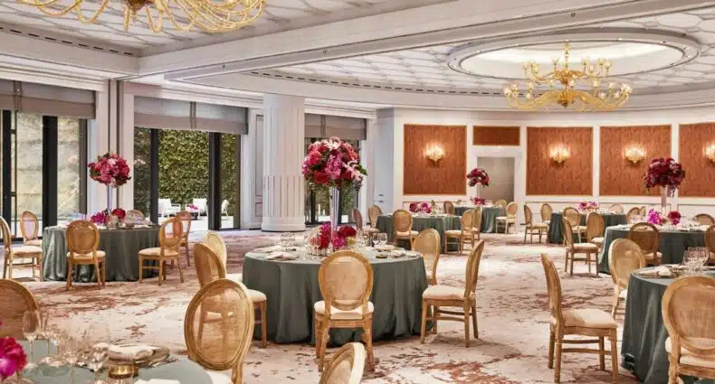 Sala de jantar do Four Seasons Hotel Madrid, uma das recomendações de hotéis de luxo em Madri. O local tem decoração luxuosa com lustres dispostos no teto e altos vasos de flores nas mesas espalhadas. As mesas redondas estão postas com conjuntos de jantas e há cadeiras ao redor. Nas paredes carmim e brancas há mais abajures e, ao fundo da sala, uma porta aberta leva à varanda com cadeiras.