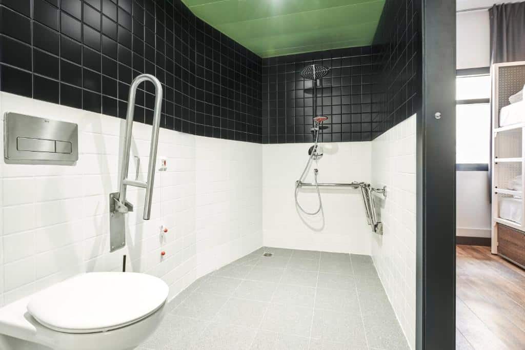 Banheiro acessível para PCD no Generator Madrid, uma das recomendações de hostels em Madri. O local é amplo e tem uma privada com barras de apoio no canto esquerdo. Ao fundo fica a área de banho com chuveiro e barras de apoio. A porta aberta fica em frente à privada e mostra um pouco do quarto com beliche.