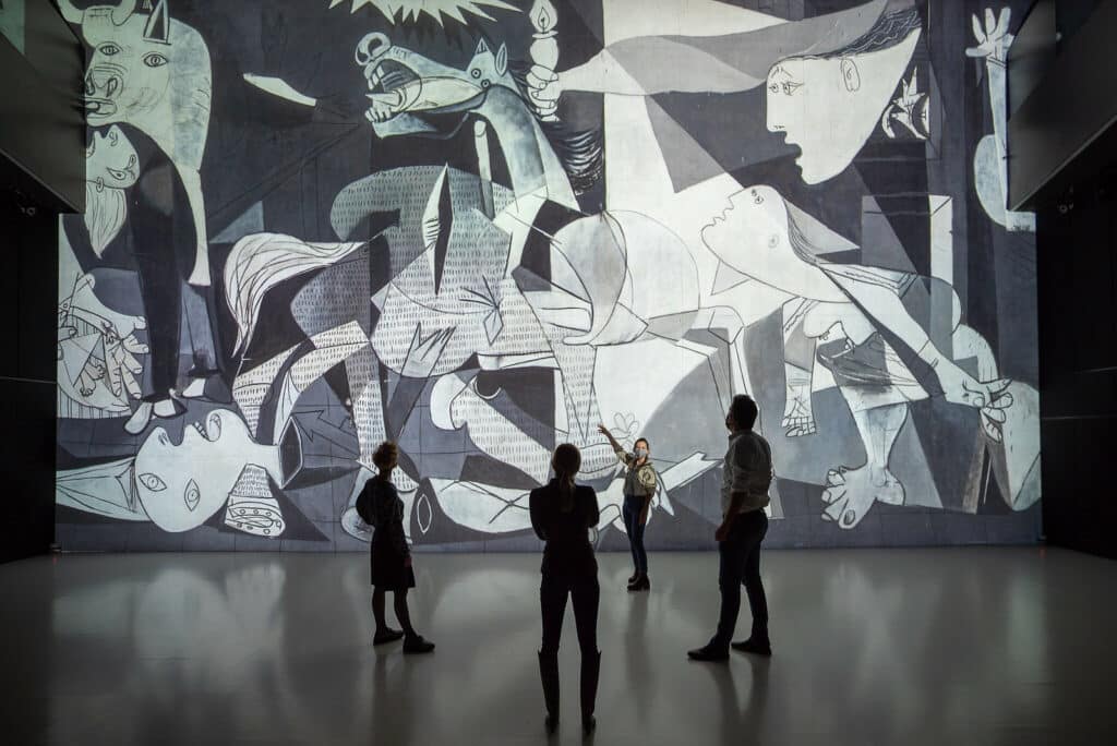 Três pessoas de costas olhando a obra Guernica, de Picasso no Museu Reina Sofía enquanto uma mulher de frente para eles aponta para a imagem como se estivesse explicando algo. - Foto: Ars Electronica via Flickr