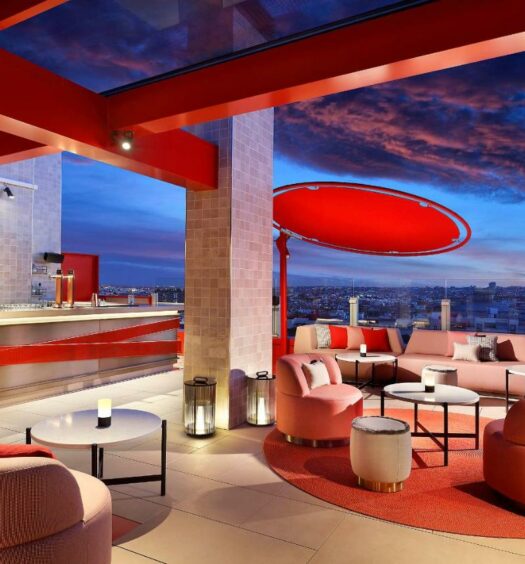 Terraço do Hard Rock Hotel Madrid, uma das recomendações de hotéis em Madri. Decorado em tons de laranja e com um lindo céu por trás, o local é repleto de sofás e poltronas. Ali há um bar ao fundo, e uma barreira de vidro protege a beirada do local que tem vista para a cidade.