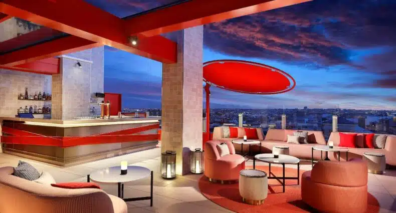 Terraço do Hard Rock Hotel Madrid, uma das recomendações de hotéis em Madri. Decorado em tons de laranja e com um lindo céu por trás, o local é repleto de sofás e poltronas. Ali há um bar ao fundo, e uma barreira de vidro protege a beirada do local que tem vista para a cidade.