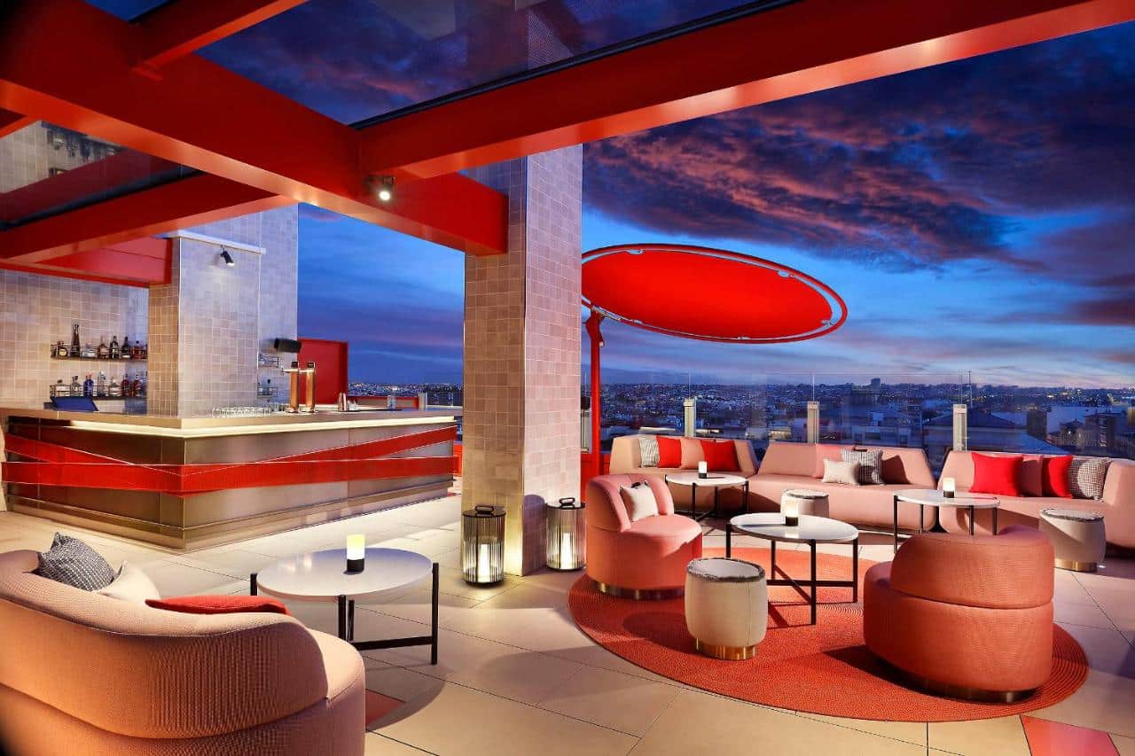 Terraço do Hard Rock Hotel Madrid, uma das recomendações de hotéis em Madri. Decorado em tons de laranja e com um lindo céu por trás, o local é repleto de sofás e poltronas. Ali há um bar ao fundo, e uma barreira de vidro protege a beirada do local que tem vista para a cidade.