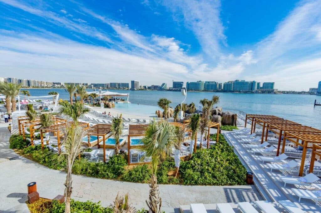 Área externa do Hilton Abu Dhabi Yas Island. Do lado direito cadeiras de tomar sol, um caminho na frente, atrás um pequeno jardim, com cadeiras de tomar sol e pequenas piscinas para limpar o pé. Atrás o mar.
