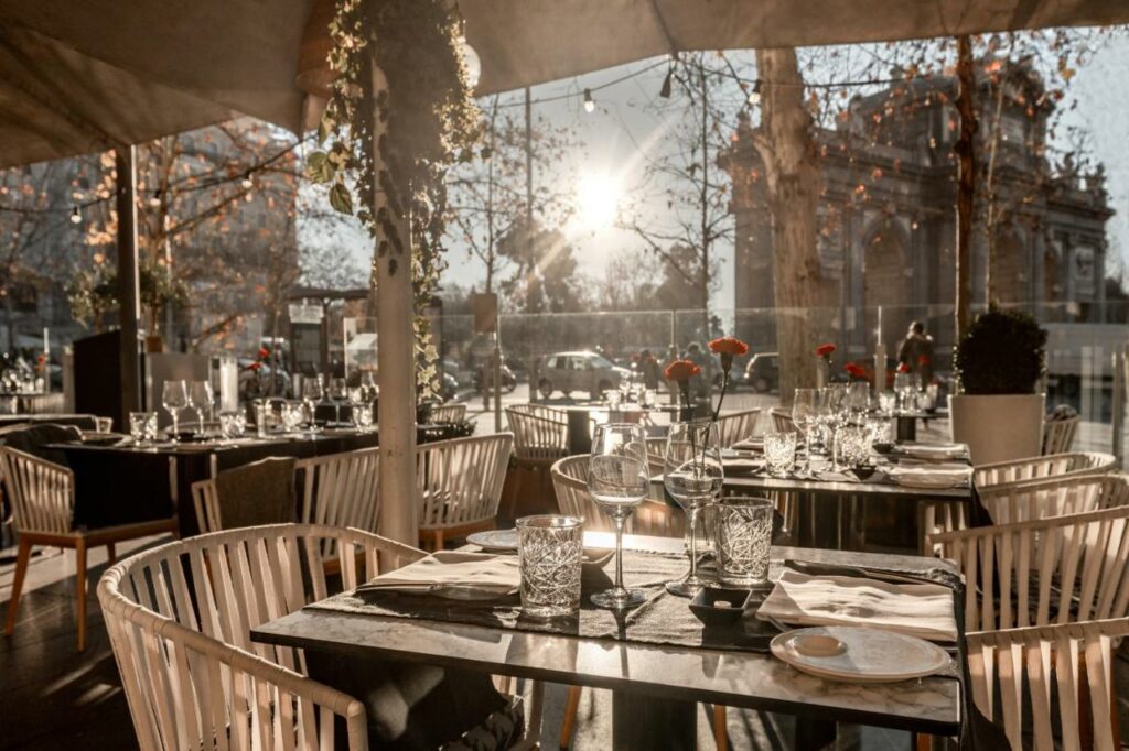 Restaurante do Hospes Puerta de Alcalá. O sol está refletindo nas paredes de vidro e iluminando o local que tem cadeiras e mesas postas de modo sofisticado. As tendas cobrem o local e tem plantinhas penduradas.