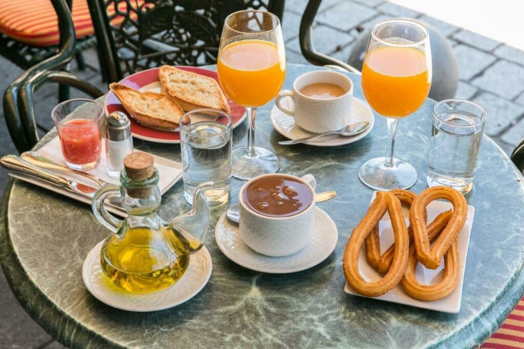 Mesa de café da manhã no Hostal Central Palace Madrid. Pratos com pães e churros estão dispostos na mesa, assim como taças de suco, copos de água e xícaras de café. Há também condimentos como azeite e saleiro.