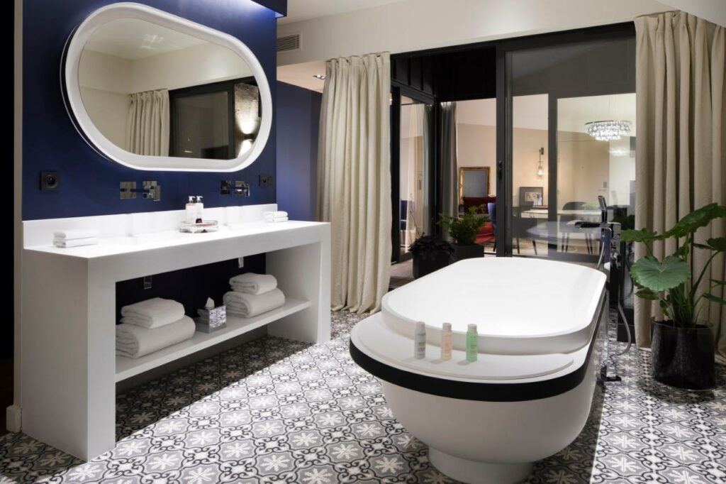Banheiro do Hôtel Cardinal Bordeaux Centre. No meio dele está uma banheira e ao lado esquerdo, encostada na parede está a pia. Acima dela está um espelho grande. Do lado direito da banheira é possível ver um vaso de planta.