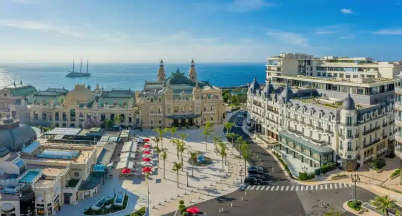 Fachada do Hôtel de Paris Monte-Carlo. Um hotel no lado esquerdo, atrás outro hotel quase no meio, uma rua na vertical e outro hotel no lado direito. Na calçada no meio, uma fonte e palmeiras. No fundo da imagem, o mar. Foto para ilustrar post sobre hotéis em Mônaco.
